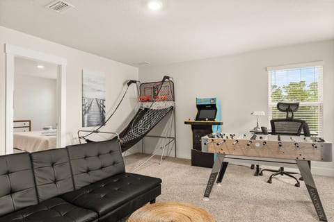 Game room featuring an indoor basketball arcade, retro-style video game machine, foosball table, and a cozy seating area with a white sofa and teal cushions. Decor includes a surfboard, wall-mounted TV, and natural light from multiple windows.