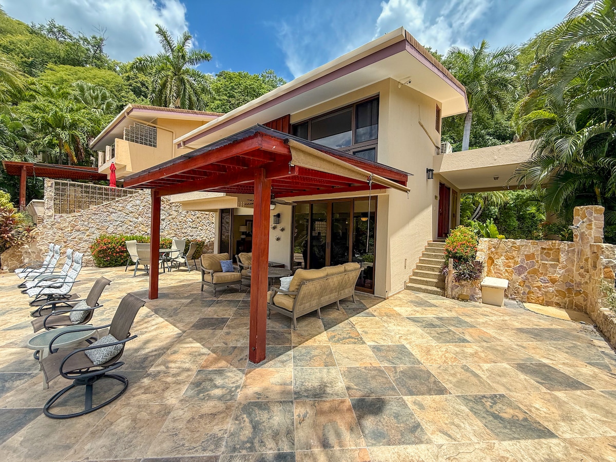 Stone-tiled patio framed by lush greenery and vibrant flowers, offering a panoramic view of the beach and Pacific horizon—perfect for morning coffee or sunset reflection in total privacy.
