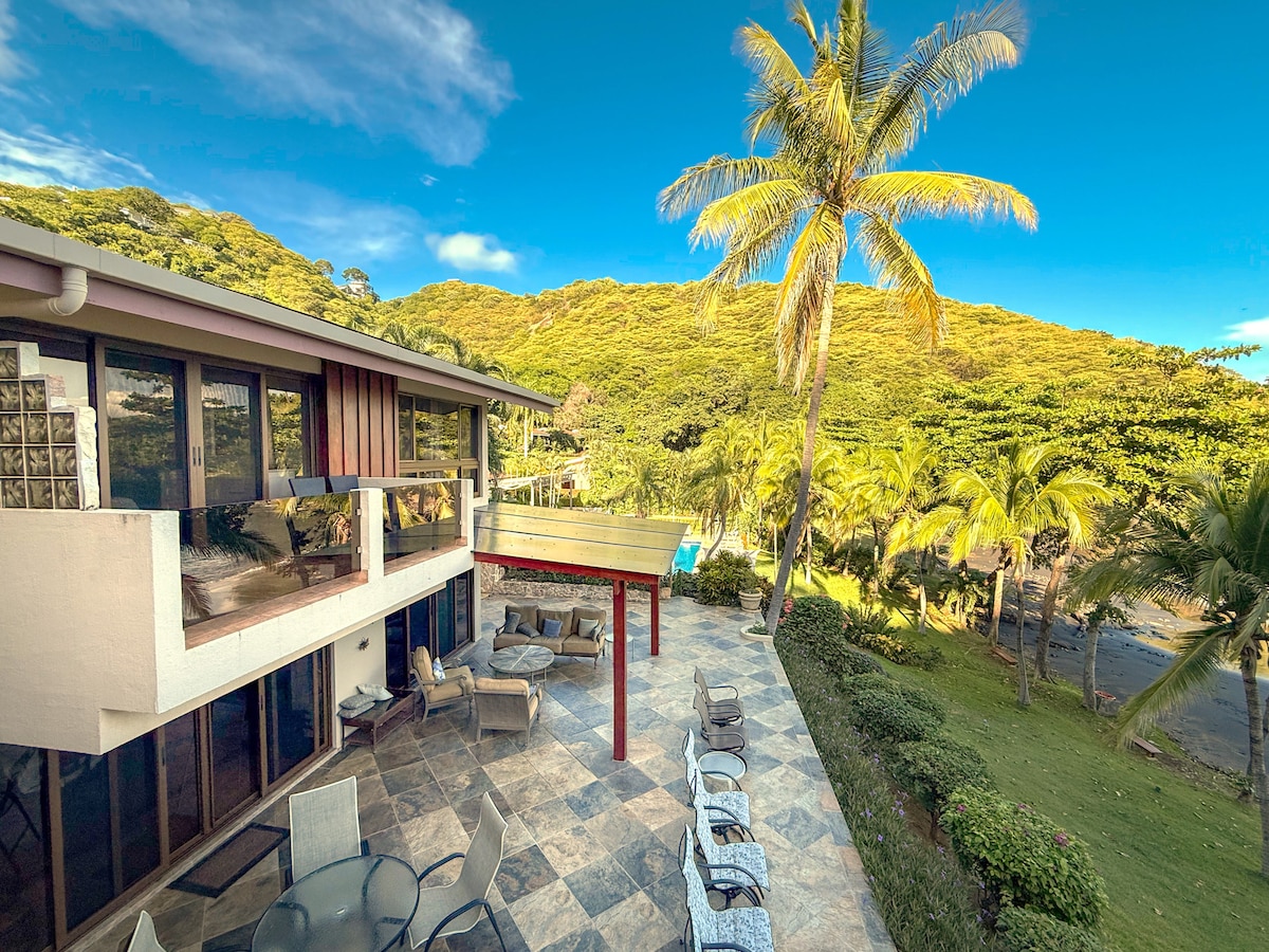 Expansive stone-tiled patio with cushioned lounge chairs, shaded dining, and lush garden views—ideal for sunrise coffee, sunset gatherings, or simply soaking in the coastal breeze.