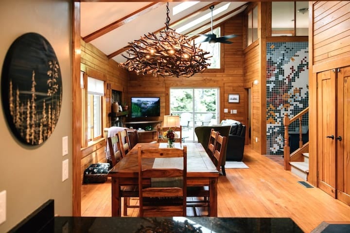 This view from the kitchen showcases an open floor plan featuring a dining table that seats six, a wood-burning stove set against a unique tiled mosaic wall, and abundant natural light from skylights, windows, and a sliding glass door.