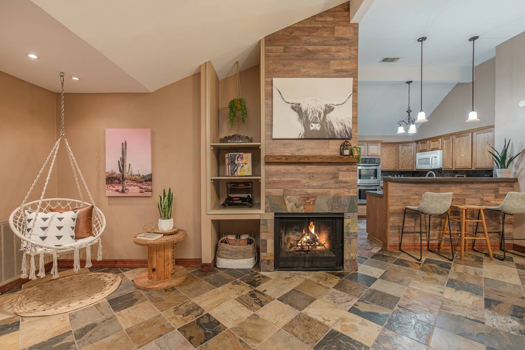 Experience comfort and charm in this living room, featuring a swing, vibrant paintings, and modern fire built into the wall, with bar stools outside the kitchen for socializing