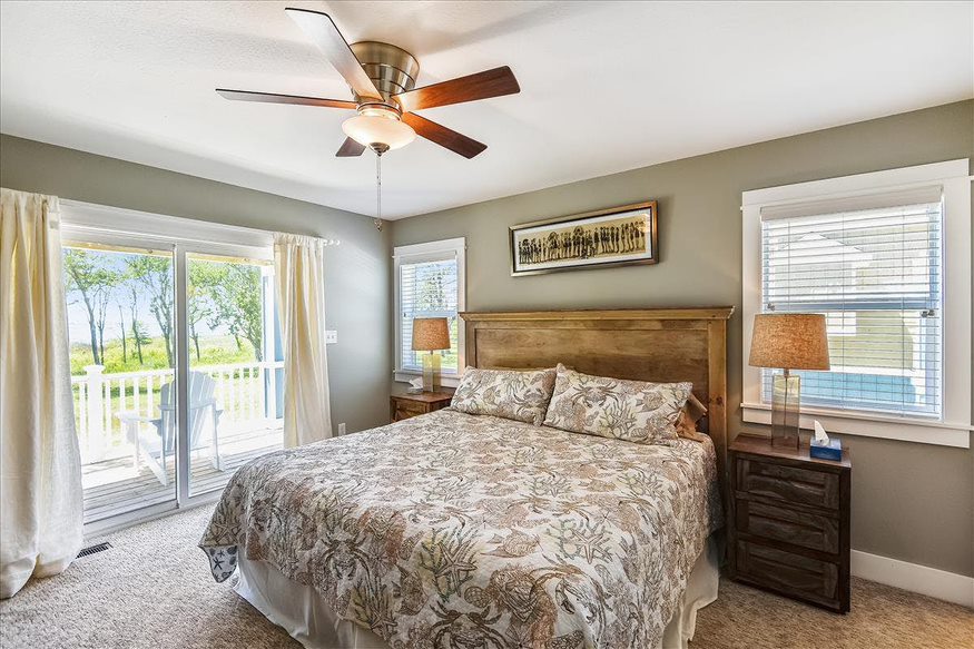 A bedroom featuring sliding doors, a ceiling fan, large windows, and a captivating picture hung above the bed.